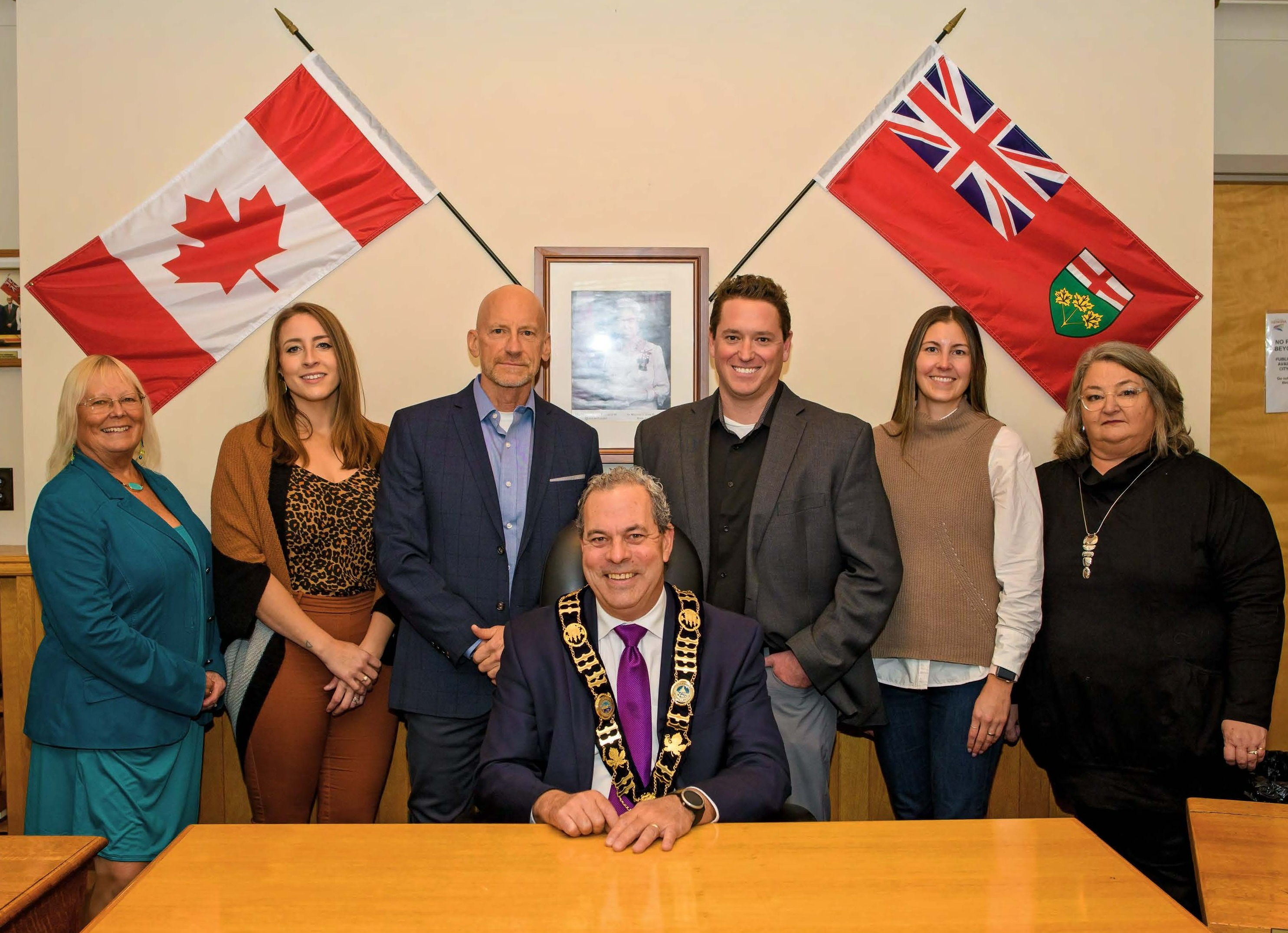 Four women and two men standing behind one man sitting at desk. The flags of Canada and Ontario are on the wall behind the group.